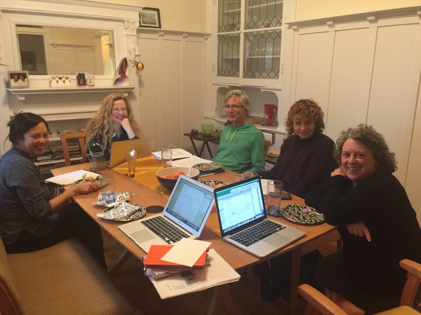 Five people seated around a table smiling, laptops and notebooks open during the first writing session for Four Chairs & a Bench.